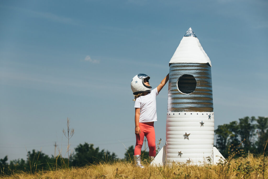 Happy child girl dressed in an astronaut costume playing with hand made rocket. Summer outdoor.